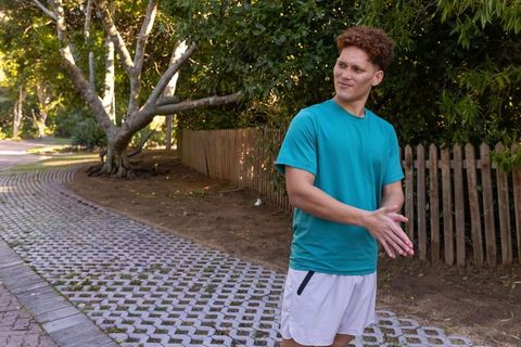 Young man standing on curved paver path by picket fence in teal shirt and shorts