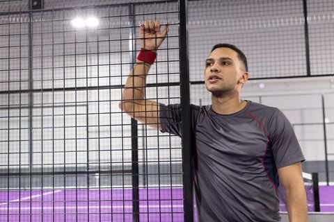 Male athlete leaning on mesh fence at purple court wearing gray shirt, red wristband