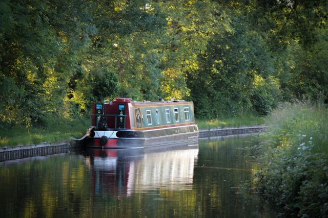 Houseboat resting on calm canal water surrounded by lush green trees and summer foliage. Ideal for concepts related to peaceful getaways, nature retreats, and outdoor adventures. Useful for travel websites, vacation brochures, or nature blogs.