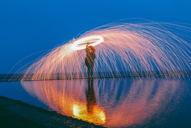 Person spinning steel wool creating trails of light and sparks by the reflective water at sunset by the sea. Ideal for themes related to creativity, night photography, light painting, and dramatic visuals.