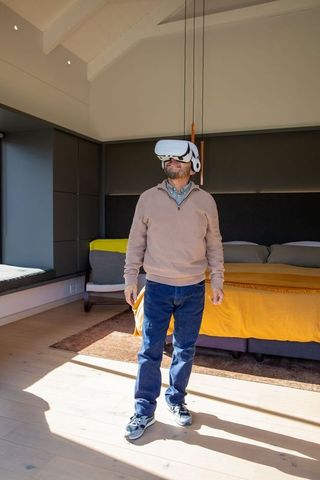 Asian man wearing vr headset standing in sunlit modern bedroom reaching toward duvet