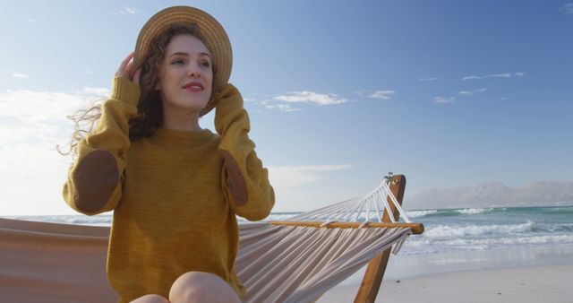 Young woman sitting on a hammock on a beach, wearing a golden sweater and sun hat, gazing into the distance. Waves gently rolling onto the shore and a clear blue sky create a peaceful atmosphere. Perfect for promoting travel, vacation retreats, leisure lifestyle, or outdoor apparel.