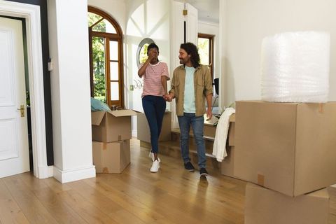 Diverse couple holding hands moving into sunlit home foyer surrounded by moving boxes