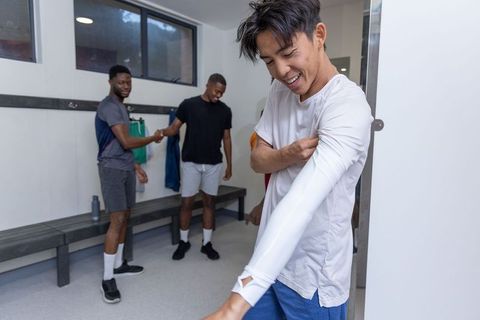 Diverse male athletes preparing in locker room, asian player pulling on white training top