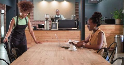 Cafe coworking scene: woman reviewing documents and working on laptop with barista background