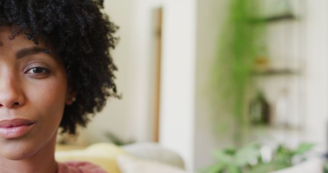 This portrait shows a young African-American woman with natural curly hair. She appears confident, captured from a close-up perspective, set indoors with blurred background emphasizing home environment. Suitable for promotional materials focusing on diversity, beauty products for natural hair, and lifestyle blogs.