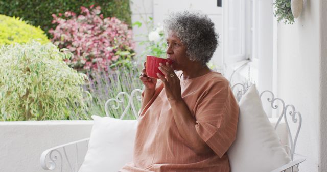A senior woman with curly hair is enjoying a cup of coffee while sitting on a white veranda with white cushions. Colorful shrubbery and greenery are visible in the background. This image is ideal for content related to retirement lifestyle, relaxation, peaceful morning routines, and mature wellness.