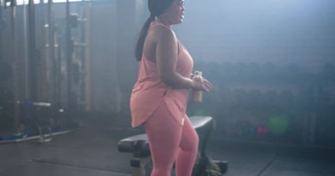 African american woman in pink activewear holding protein shaker standing by gym bench
