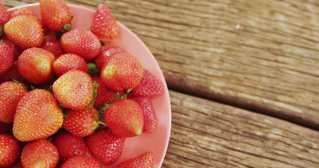 Fresh, ripe strawberries are displayed in a pink bowl set on a rustic wooden table. This colorful and vibrant image can be used for food blogs, healthy eating promotions, and summer recipe websites. The close-up view emphasizes the natural texture and juiciness of the strawberries, making it suitable for advertisements for organic produce or farm markets.