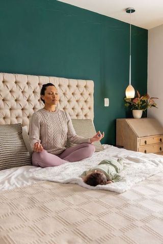 Mother meditating on bed while infant sleeping nearby in cozy bedroom