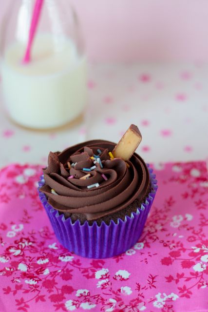 Chocolate cupcake topped with creamy frosting and sprinkles, perfect for celebrations and dessert ideas. The pink floral cloth and milk bottle add a nostalgic and inviting feel, suitable for bakery promotions, recipe blogs, and festive event themes.