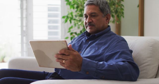 Senior man sitting comfortably on couch using tablet in a modern living room. He is concentrated and relaxed, wearing a blue shirt and enjoying his time with the digital device. Perfect for lifestyle, technology, or retirement-themed projects showcasing older adults engaging with modern technology in a home setting.