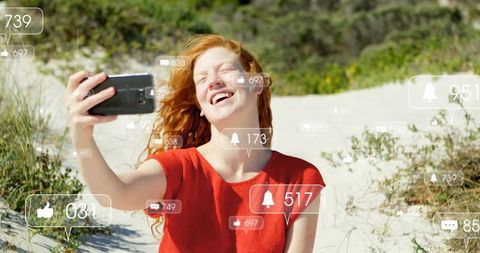 Young woman taking joyful selfie on coastal dune with social media likes and notifications