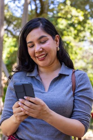 Smiling woman checking smartphone in sunny park wearing gray top and gold hoop earrings