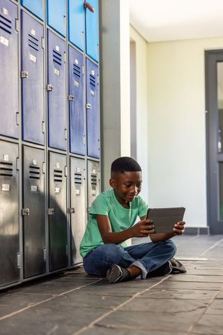 African american boy sitting in school hallway using tablet by blue lockers