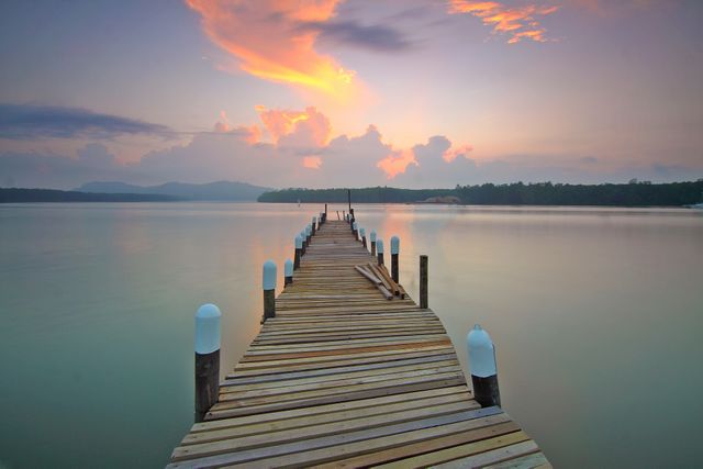 Depicts a tranquil scene with a wooden pier extending into the calm waters of a lake during a beautiful sunset. The sky features dramatic clouds with hues of orange and purple. The serene atmosphere and scenic nature make this ideal for use in travel marketing, nature posters, motivational quotes, and relaxation-themed projects.