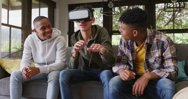 Three teenage boys bonding over a virtual reality video game in a casual living room setting. One boy is wearing a VR headset while the other two laugh and guide him. Perfect for illustrating the fun and interactive nature of modern digital entertainment, as well as friendship and youthful enjoyment of technology.