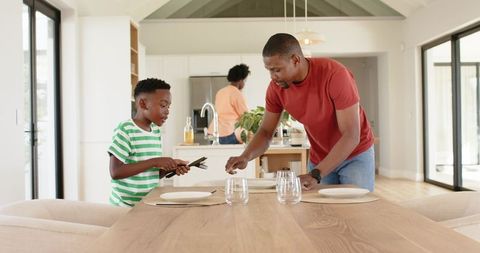 Family placing tableware in sunlit modern open-plan kitchen, father and son preparing meal