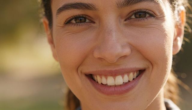 Smiling hispanic woman revealing teeth in sunlit park closeup showing brown eyes and natural smile