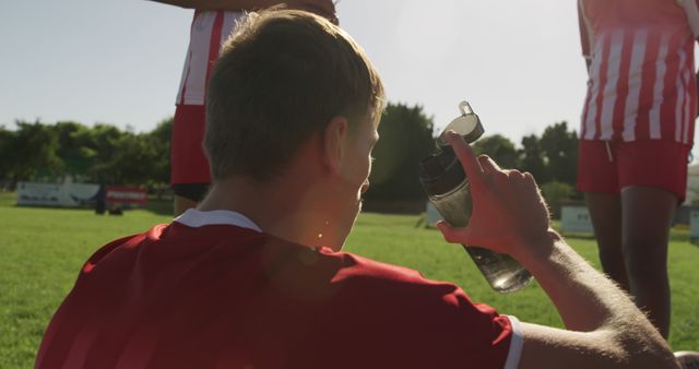 Young soccer player taking a drink from a water bottle during a break in a game on a sunlit field. Ideal for content related to youth sports, hydration, teamwork, outdoor activities, athletic lifestyle, and sportsmanship.