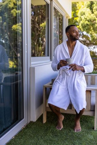 Mature black man relaxing on balcony in white robe holding coffee mug morning calm