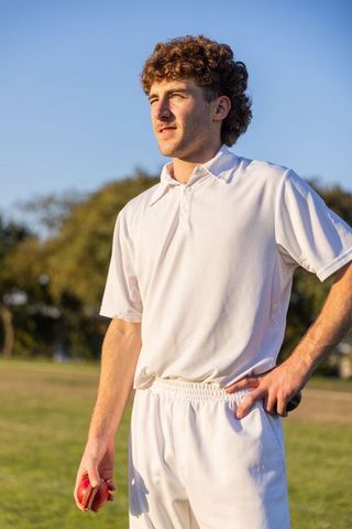 Sunlit cricketer standing on outfield holding red ball wearing white kit at golden hour