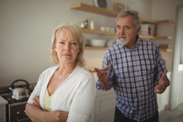 Senior couple in modern kitchen, woman with arms crossed while man in background gesturing. Ideal for content on relationship advice, family dynamics, aging, or marriage counseling.
