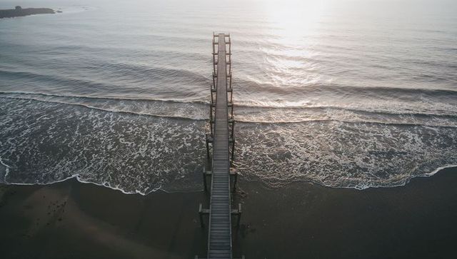 Sunlit wooden pier leading into calm ocean at dawn minimalist aerial coastal view