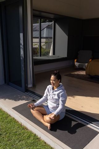 Asian woman meditating on doorstep yoga mat wearing hoodie morning sunlight serenity calm
