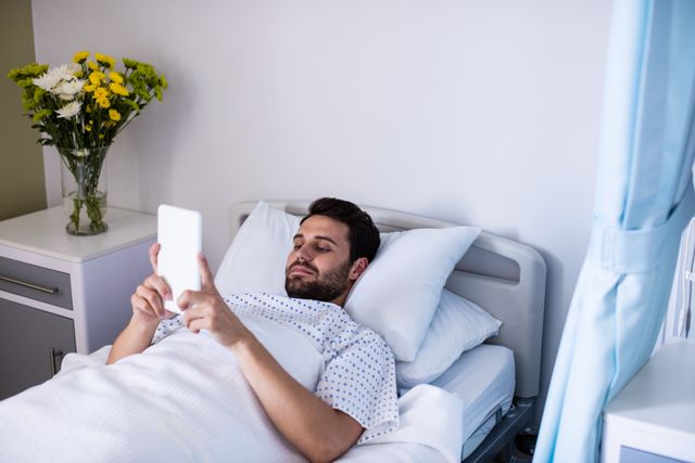 Male patient lying in hospital bed using digital tablet. Flowers on bedside table add a touch of comfort. Ideal for illustrating modern healthcare, patient care, and technology in medical settings. Useful for healthcare websites, medical brochures, and technology in healthcare articles.
