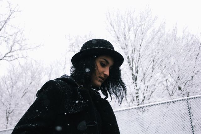 This image captures a woman wearing a black hat walking in a snowy park. Bare trees covered in snow frame the background, creating a peaceful and serene winter scene. Perfect for concepts related to winter fashion, solitude, contemplation, and the beauty of nature in cold seasons.