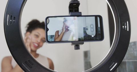 African american woman applying skincare while filming with ring light and smartphone
