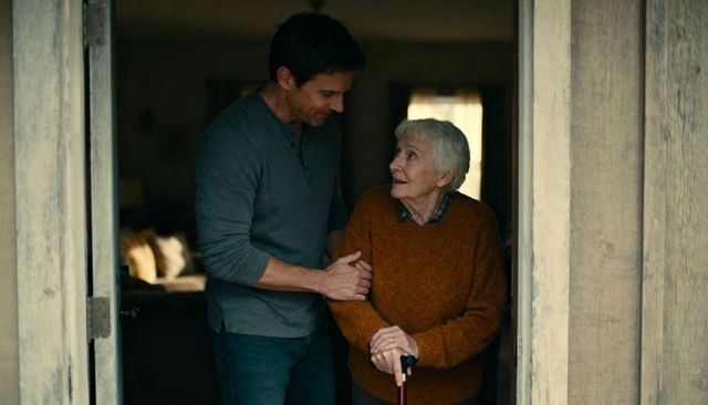 Adult son helping elderly woman with cane in rustic doorway warm caregiving moment
