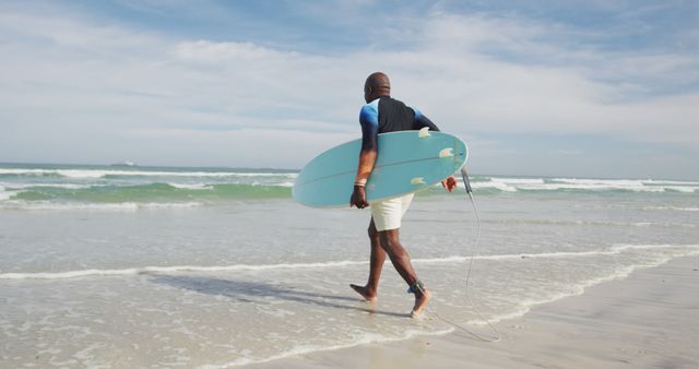 African american senior man walking on a beach holding surfboard and running into the sea. healthy outdoor leisure time.