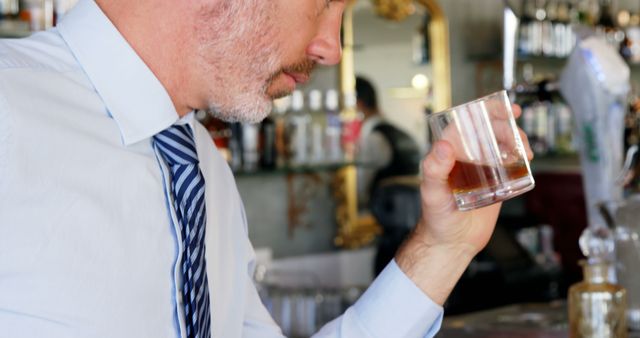 A middle-aged Caucasian businessman enjoys a glass of whiskey at a bar, with copy space. His attire and the setting suggest a moment of relaxation after a workday.