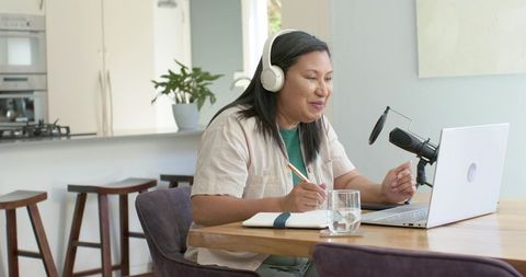 Woman recording podcast at kitchen table wearing headphones and taking notes on laptop
