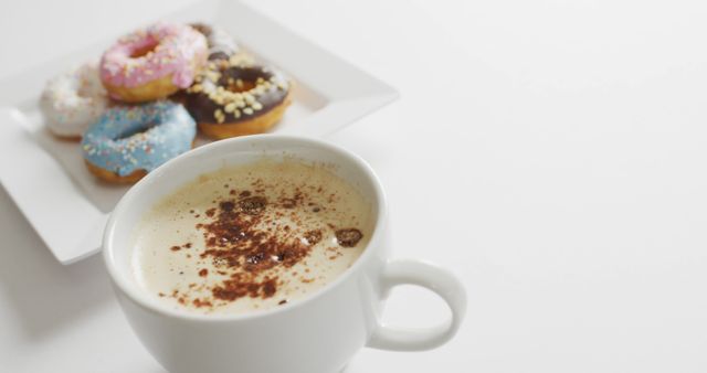 Foamy cup of coffee sprinkled with chocolate powder close-up in front of plate with assorted donuts topped with colorful icing and sprinkles against white backdrop. Ideal for breakfast spot, bakery advertisement, coworking space, and morning treat promotions.