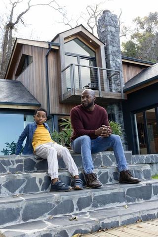 African american father and son sitting on stone steps outside modern wood and stone home