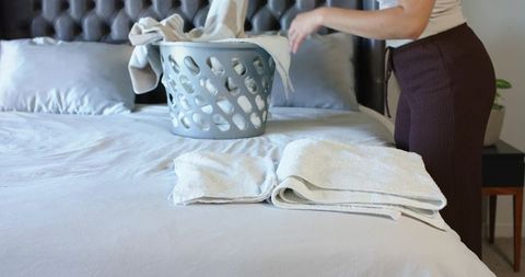 Woman arranging towels on bed with laundry basket in modern bedroom