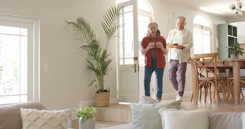 Diverse senior couple entering sunlit home carrying drinks and tray, warm hospitality
