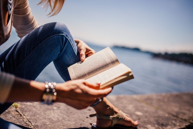 This stock photo depicts a woman reading a book by the lake on a sunny day. The individual is dressed in casual summer attire, including jeans and sandals, capturing a moment of relaxation and tranquility in nature. This image is suitable for use in articles or advertisements related to leisure, travel, outdoor activities, lifestyle, mental health, or promoting reading and education.