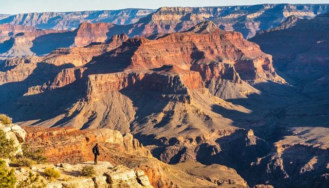 Lone hiker surveying vast red canyon from cliff ledge at sunrise light