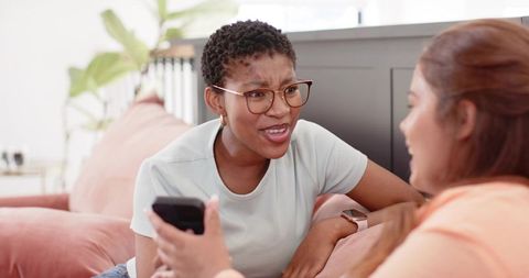 Cozy living room conversation between friends sharing smartphone and laughing on pink sofa