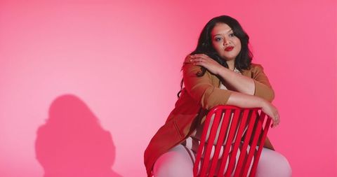 Mid adult woman leaning over red chair wearing blazer against pink gradient backdrop