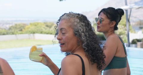 Mature woman sipping citrus cocktail poolside while friend wearing sunglasses is chatting