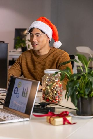 Asian man wearing santa hat working at festive office desk with laptop, lights, gift