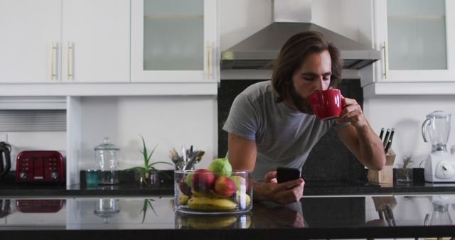 Young man drinking coffee while using smartphone in a modern kitchen. The scene includes contemporary kitchen appliances and a bowl of fruits on the counter. Useful for illustrating lifestyle, morning routine, modern living, technology use at home, and kitchen interior design.