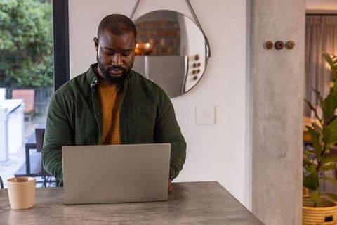 African american man working on laptop at modern kitchen island with coffee mug, focused