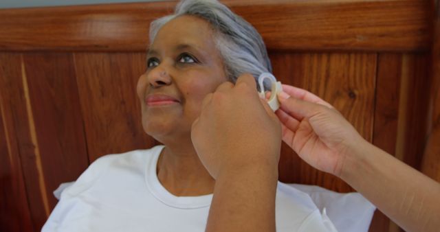 Female doctor inserting hearing aid in senior black woman ear in a comfortable home. She is smiling and relaxing on bed 4k