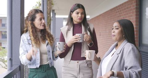 Diverse female coworkers networking on office balcony holding coffee mugs